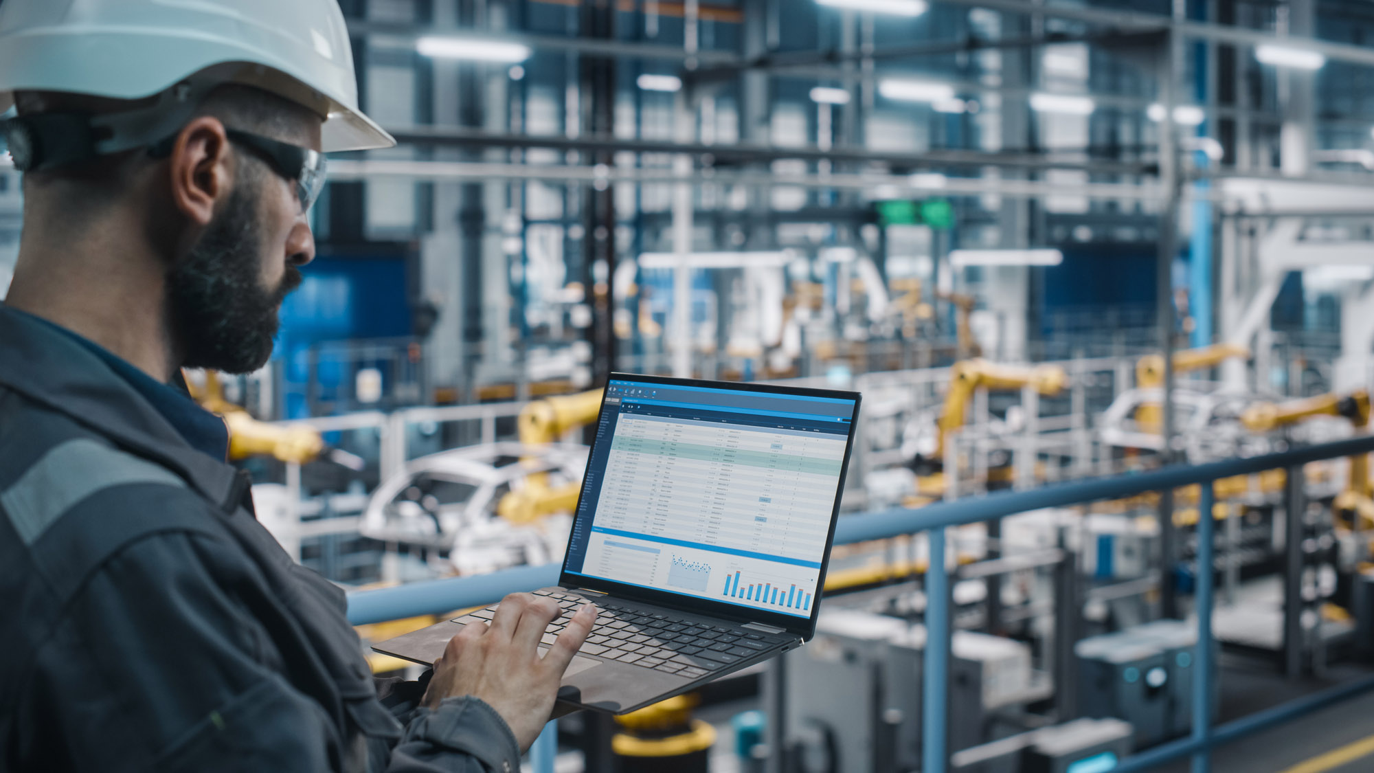 Car Factory Engineer in Work Uniform Using Laptop Computer with Spreadsheet Software. Working with Software at Automotive Industrial Manufacturing Facility Dedicated for Vehicle Production.